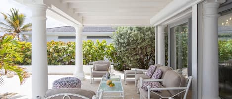 A covered outdoor seating area with white furniture, a glass table, and a view of the garden and house in the background.