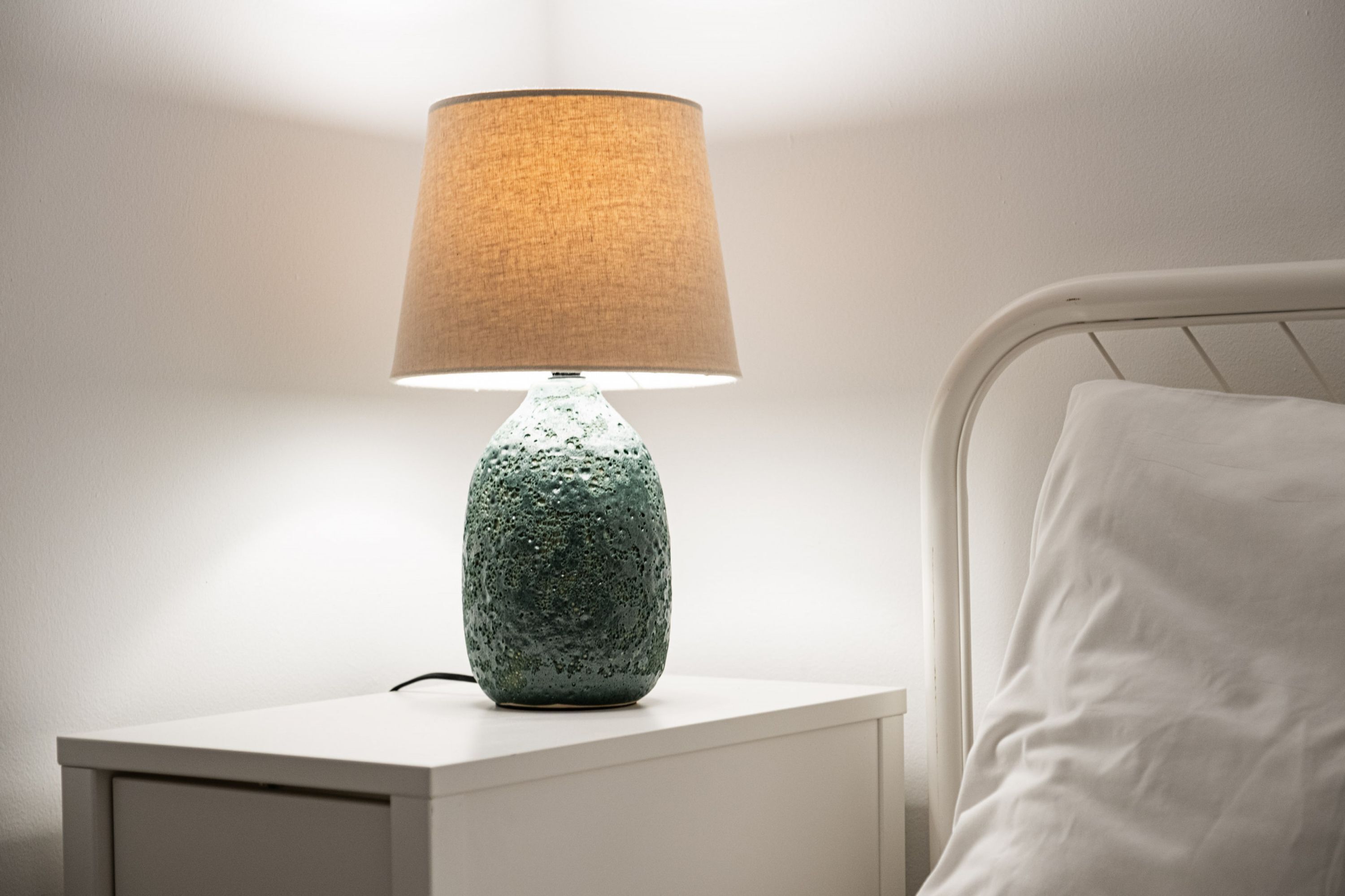 A white bedside table with a textured green lamp and a white pillow.