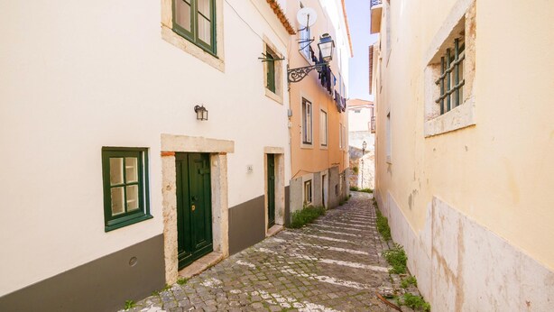 A narrow cobblestone street flanked by buildings with white and pastel-colored facades, green shutters, and a small overgrown area at the street's edge. - Nomad s Castel Lisbon - 1bdr The Reader (Lisbon)