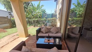 A covered outdoor seating area with a sofa and a coffee table, surrounded by greenery and a building in the background. - Luxury Flatlet in the heart of Umhlanga (uMhlanga)