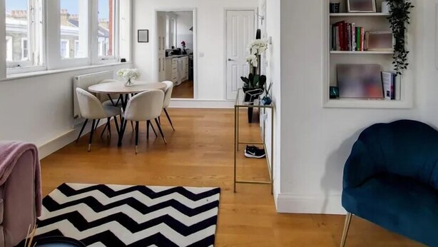 A modern living room with a blue armchair, a dining area with a round table, and a bookshelf with books and a plant. - Apartment Sleeps 2 with Free Parking (London)