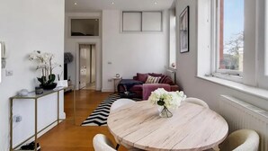 A modern living room with a round wooden table, a sofa, and a window with a view. - Apartment Sleeps 2 with Free Parking (London)