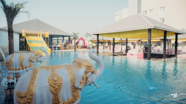 Ein Schwimmbecken mit dekorativen Elefantenstatuen, ein Kinderspielbereich und eine überdachte Sitzgelegenheit mit Sonnenschirmen.