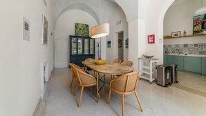 A dining area with a round table, six wicker chairs, a central light fixture, and a kitchen area with a tiled backsplash and green cabinets.