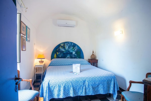 A bedroom with a bed, a chair, a nightstand, a lamp, and a wall-mounted air conditioner.