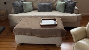 Ein Wohnzimmer mit einem beigen Sofa, einem kleinen Hocker und einem Fenster mit Blick auf ein Gebäude.
