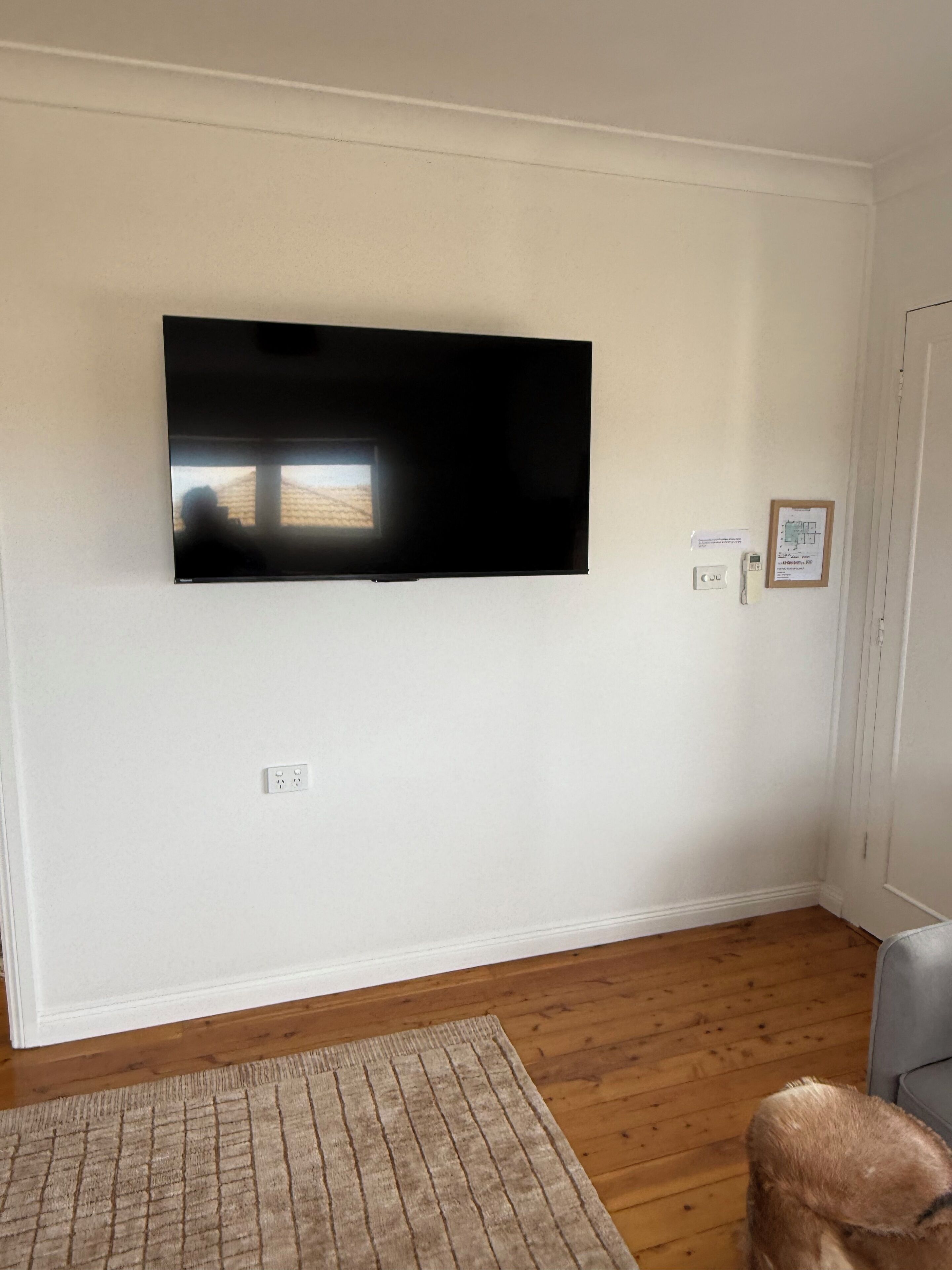 Una habitación con televisión montada, suelo de madera, sofá gris y una alfombra pequeña.