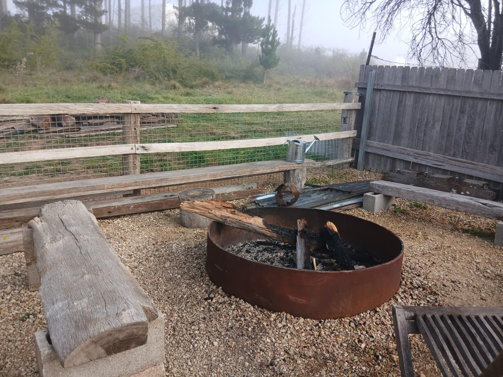 A rustic outdoor area with a metal fire pit, a wooden bench, and a gravel ground.