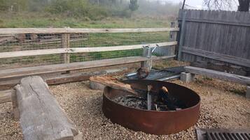 A rustic outdoor area with a metal fire pit, a wooden bench, and a gravel ground.