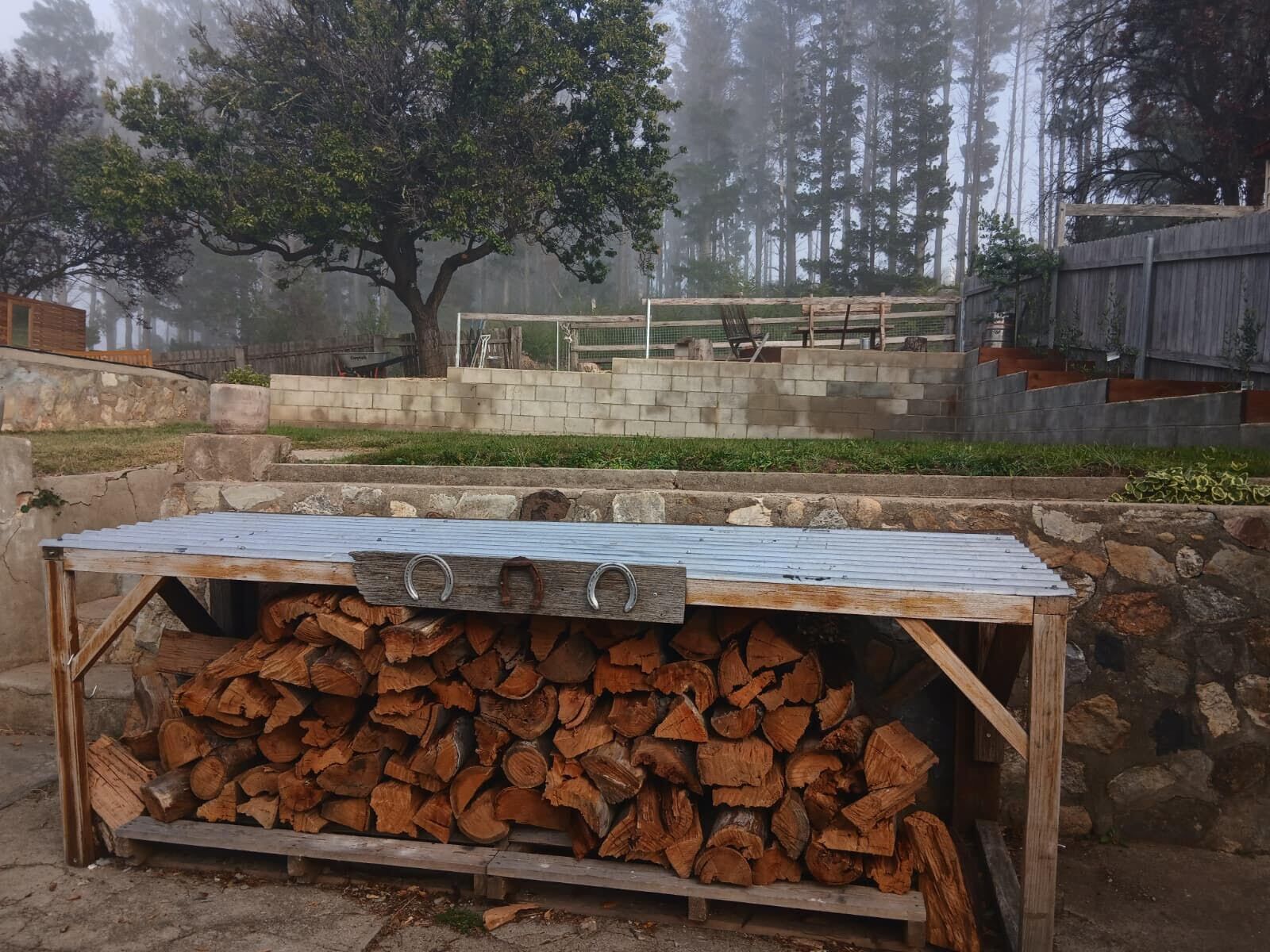 A rustic outdoor grill area with a stack of firewood, a stone wall, and a view of a forested area.