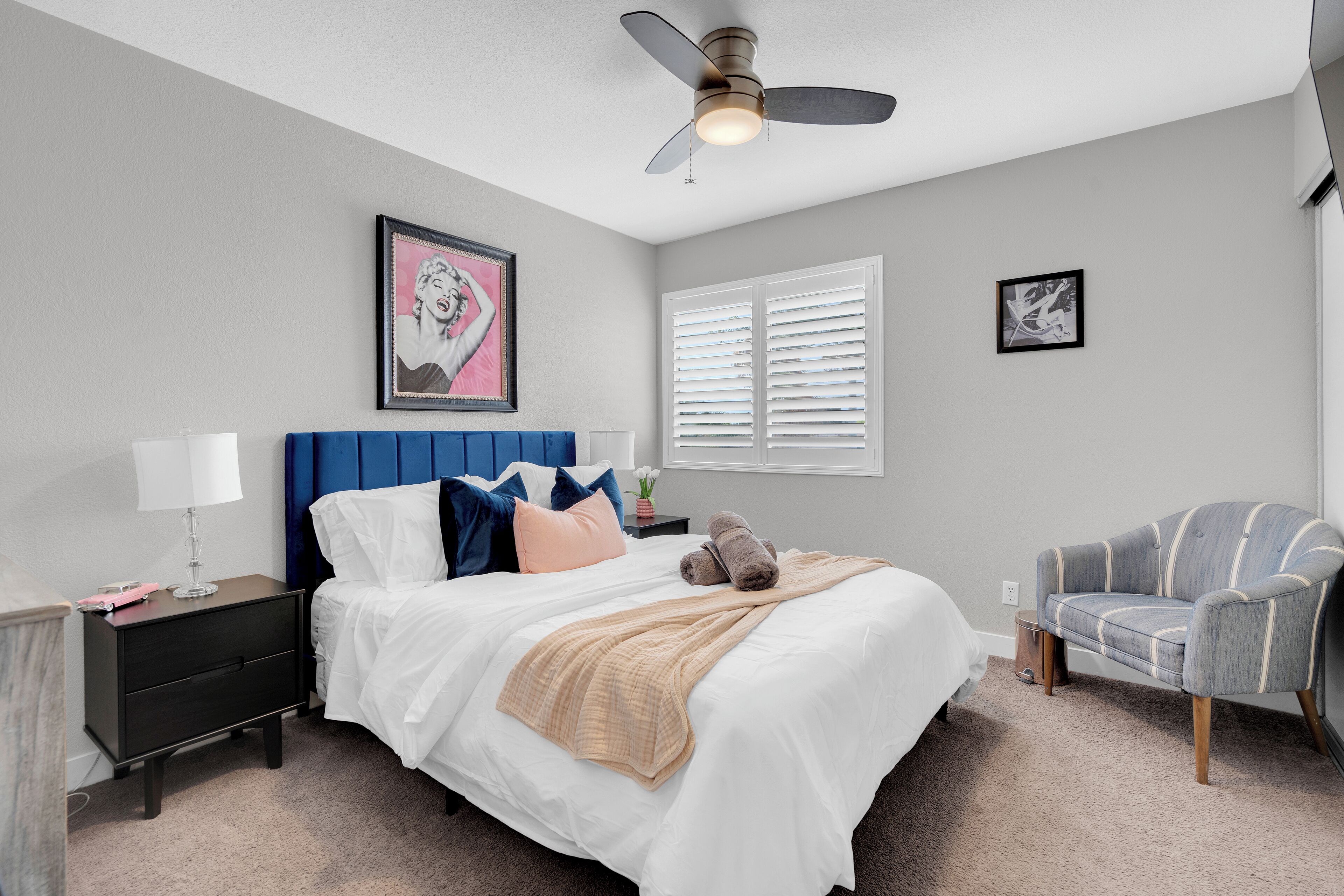 A bedroom with a bed, nightstand, chair, ceiling fan, and artwork on the walls.