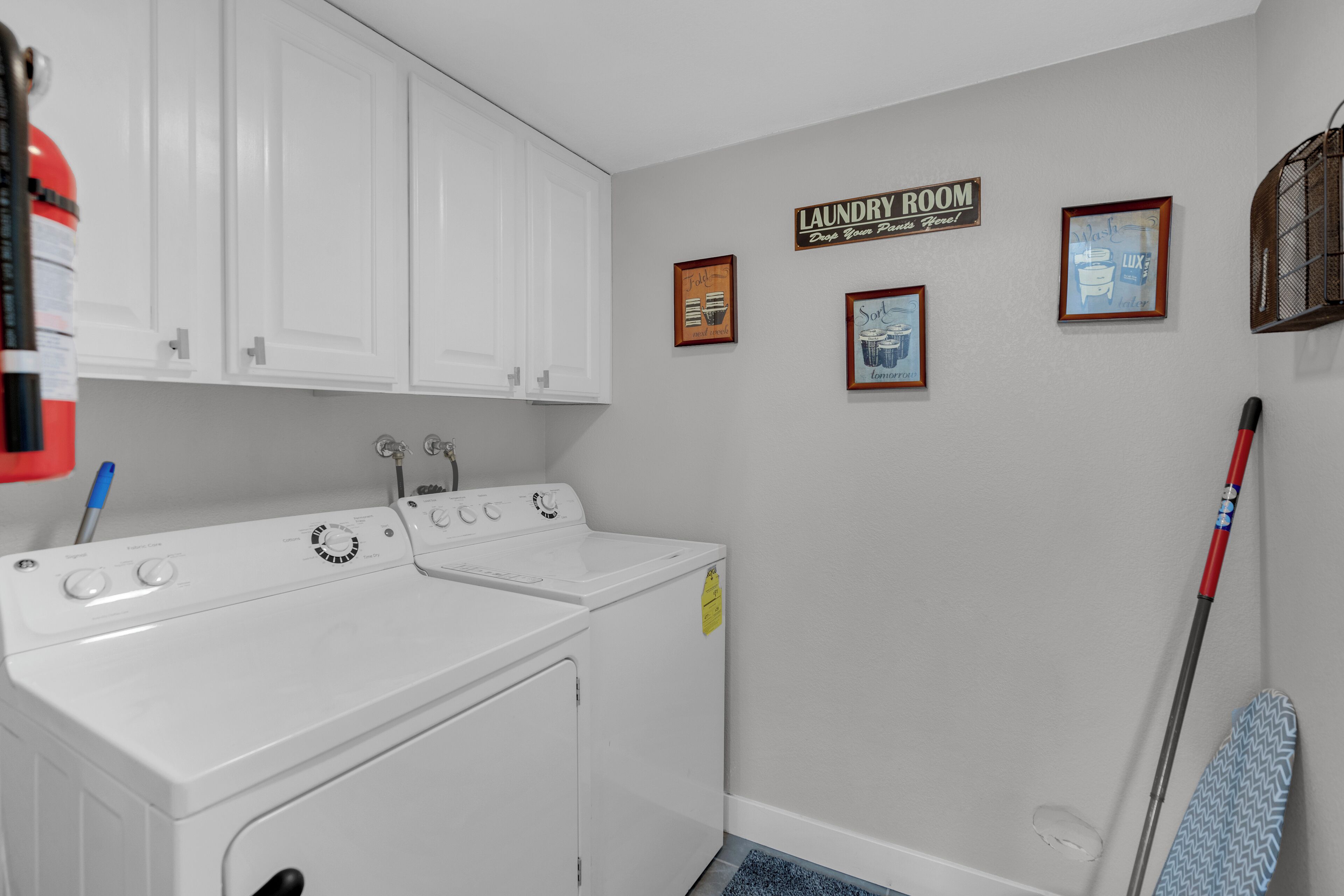A laundry room with a washing machine, dryer, and ironing board. There are framed pictures on the wall and a mop leaning against the wall.
