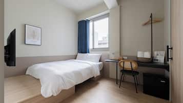 Habitación de hotel compacta con una cama individual, un escritorio con silla, una mesita con cesta, un cesto de basura negro y una ventana con cortinas.
