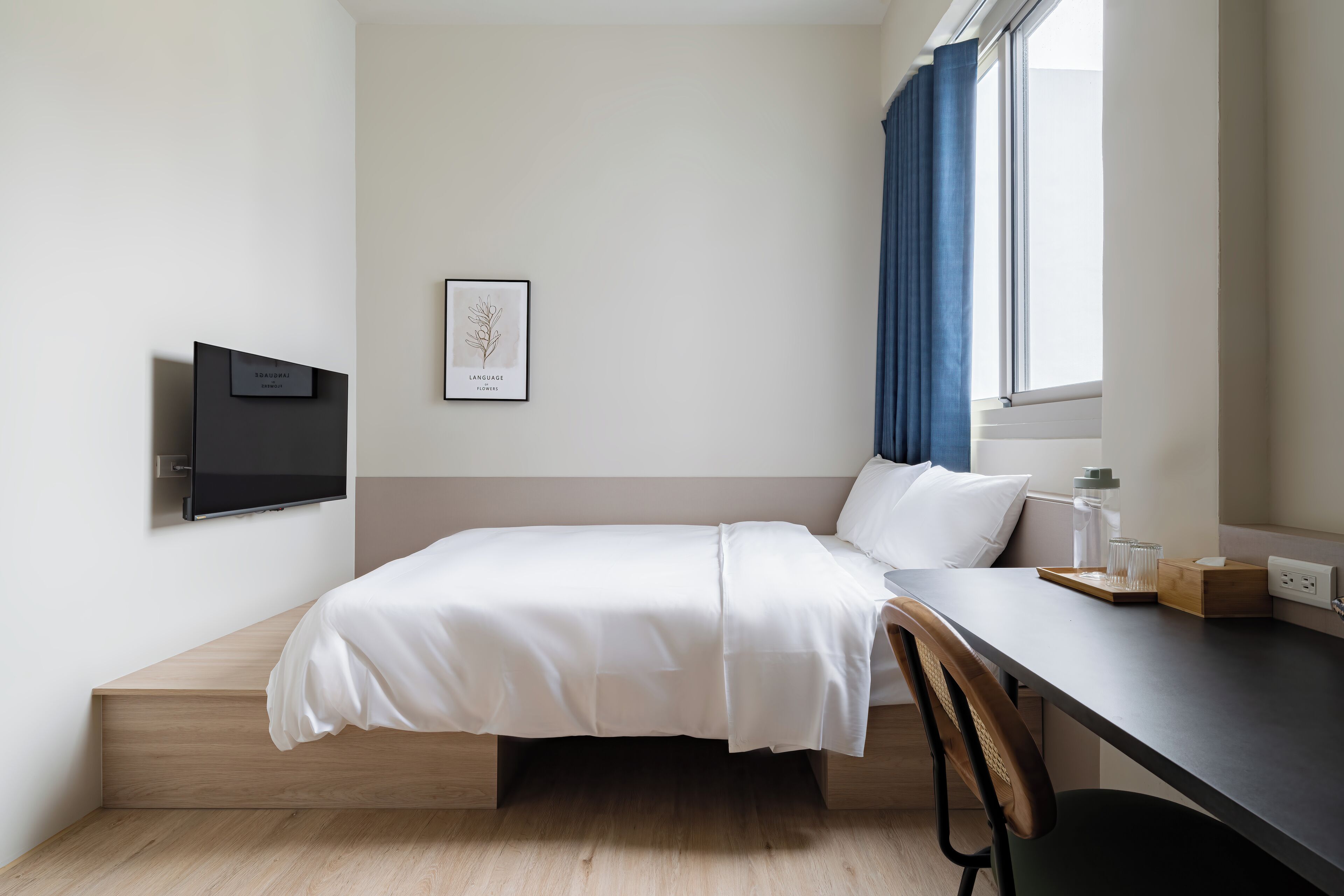 A minimalist hotel room with a bed, a desk, a chair, a TV, and a window with curtains.