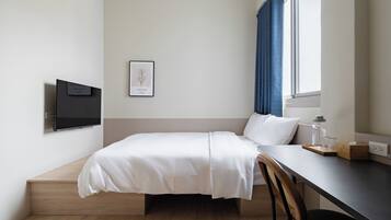 A minimalist hotel room with a bed, a desk, a chair, a TV, and a window with curtains.