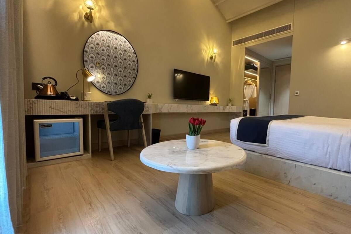 Ein kleines Hotelzimmer mit einem Bett, einem Schreibtisch mit Computer, einem Sessel, einem runden Tisch mit Blumenvase und einem an der Wand montierten Fernseher.