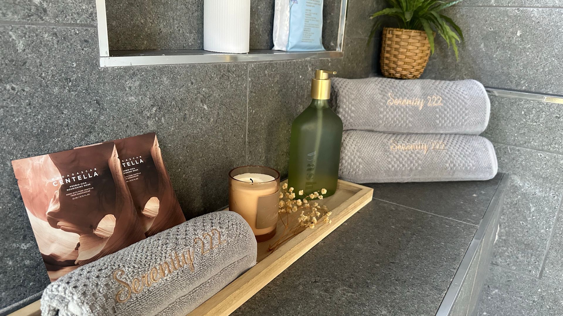 Un espace spa avec une étagère en bois sur laquelle sont posés une bouteille, des bougies et une serviette brodée du motif « Serenity 222 ».