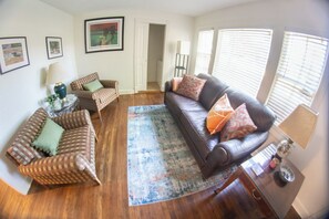 A living room with a brown leather sofa, a brown checkered sofa, and a glass coffee table. There are framed pictures on the wall, a lamp, and a window with blinds. - The Blue Jay House in North Fondren (Jackson)