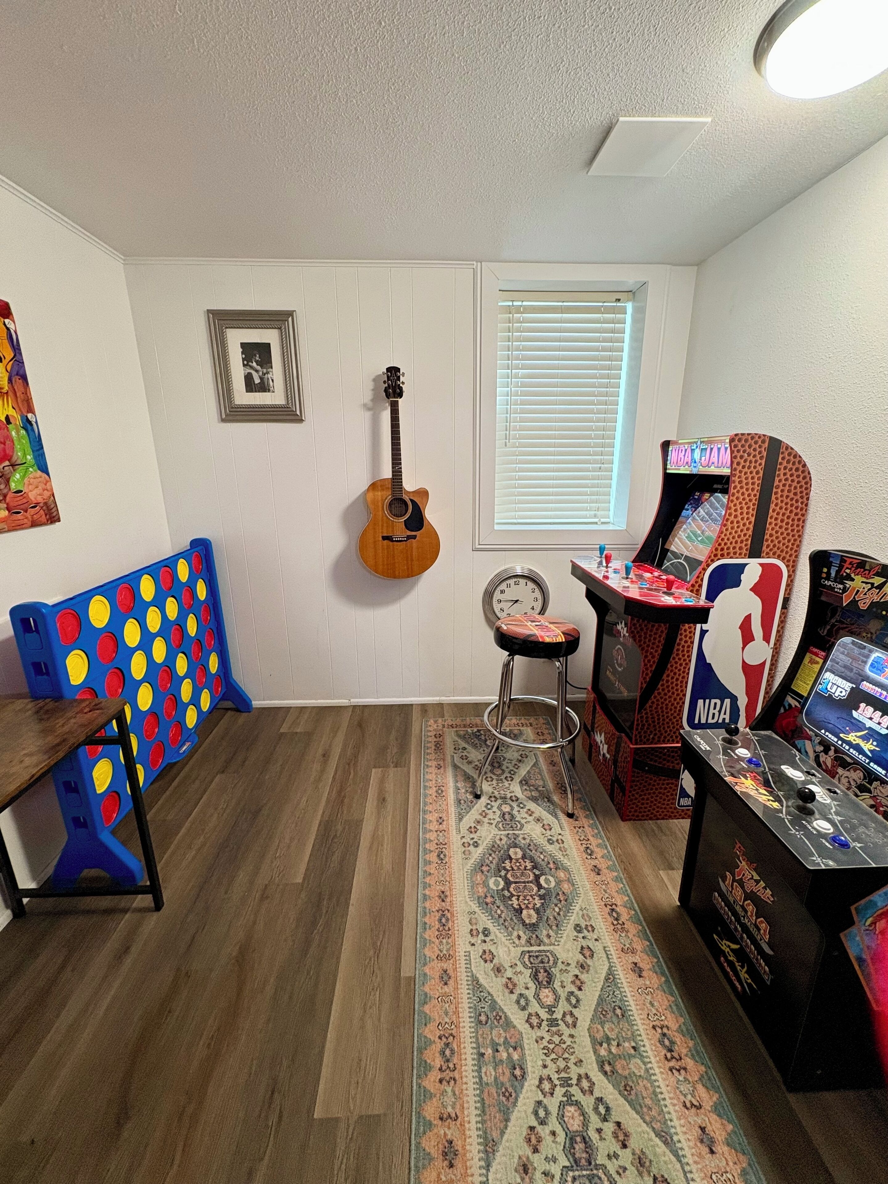 Una habitación con piso de madera, una mesa de juegos azul y amarilla, una guitarra, un reloj y una máquina de videojuegos con temática de la NBA.