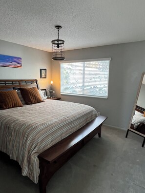 Une chambre à coucher avec un grand lit, une table de chevet, une commode, un luminaire suspendu, une fenêtre avec des stores et un tableau accroché au mur.