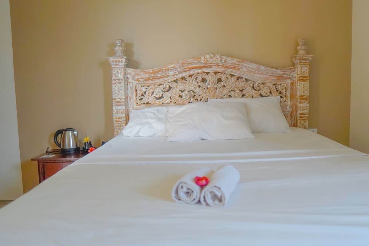 Una cama bien hecha con un cabecero decorativo, una toalla enrollada con una flor roja y una tetera sobre una mesita de noche.