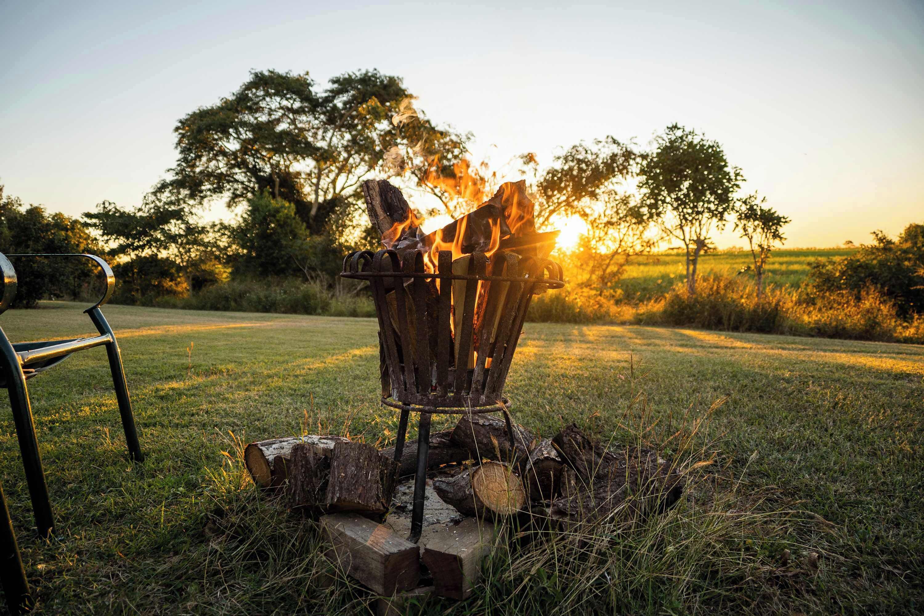 Un feu de camp avec des bûches et une chaise en métal, dans un champ herbeux avec des arbres en arrière-plan.