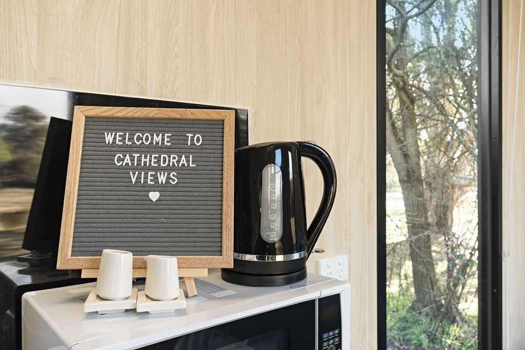 A sign reading "WELCOME TO CATHEDRAL VIEWS" on a blackboard, a black coffee maker, and two white cups on a counter.
