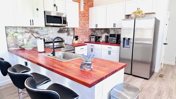 A modern kitchen with white cabinets, a wooden island with bar stools, stainless steel appliances, and a brick accent wall. - 4bed/3bath House in St. Louis (St. Louis)