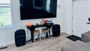 A living room with a large flat-screen TV mounted on the wall, a black speaker on the left, and a smaller speaker on the right. There are two wooden stools in front of a black entertainment unit with various items on it. A white door is visible on the right side of the image. - 4bed/3bath House in St. Louis (St. Louis)