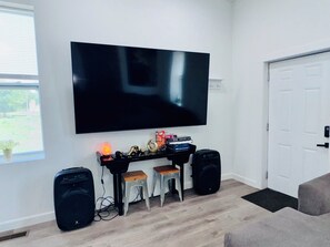 Un salon avec un grand téléviseur à écran plat fixé au mur, un haut-parleur noir à gauche et un haut-parleur plus petit à droite. On peut voir deux tabourets en bois devant un meuble noir contenant divers objets. Une porte blanche est visible sur le côté droit de l’image.