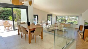 A modern living room with a glass partition, a dining area with a wooden table and chairs, and a view of a swimming pool and greenery outside. - Villa 6 rooms - 10 people (8 adults max) - Private pool - Air conditioning - WiFi - Sainte Maxime (STE MAXIME)