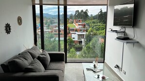 A modern living room with a grey sofa, a glass coffee table, and a large window offering a view of a residential area with houses and greenery. - Apartamento Moderno en Ciudad Cayalá (Ciudad de Guatemala)