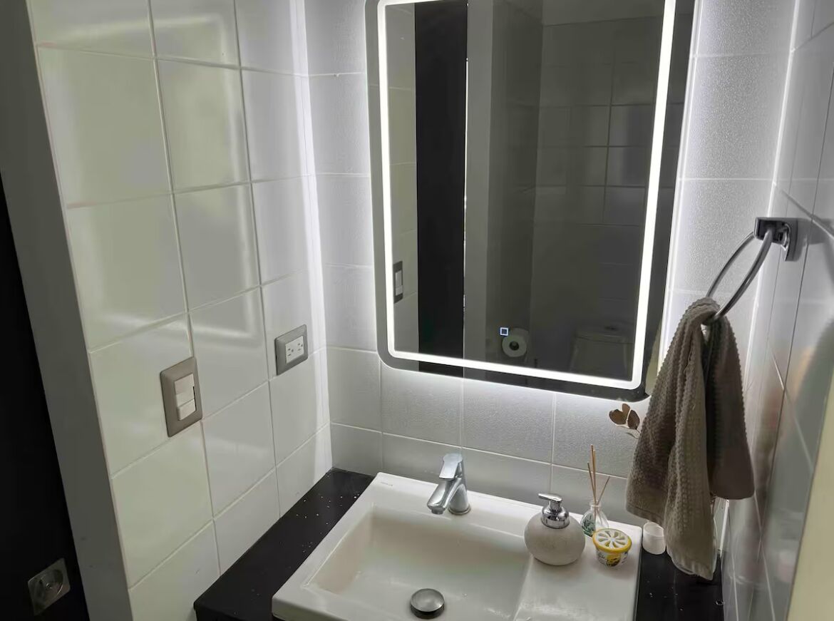 Une salle de bain avec des murs recouverts de carreaux blancs, un comptoir noir, un lavabo avec un robinet, un miroir et un porte-serviettes.
