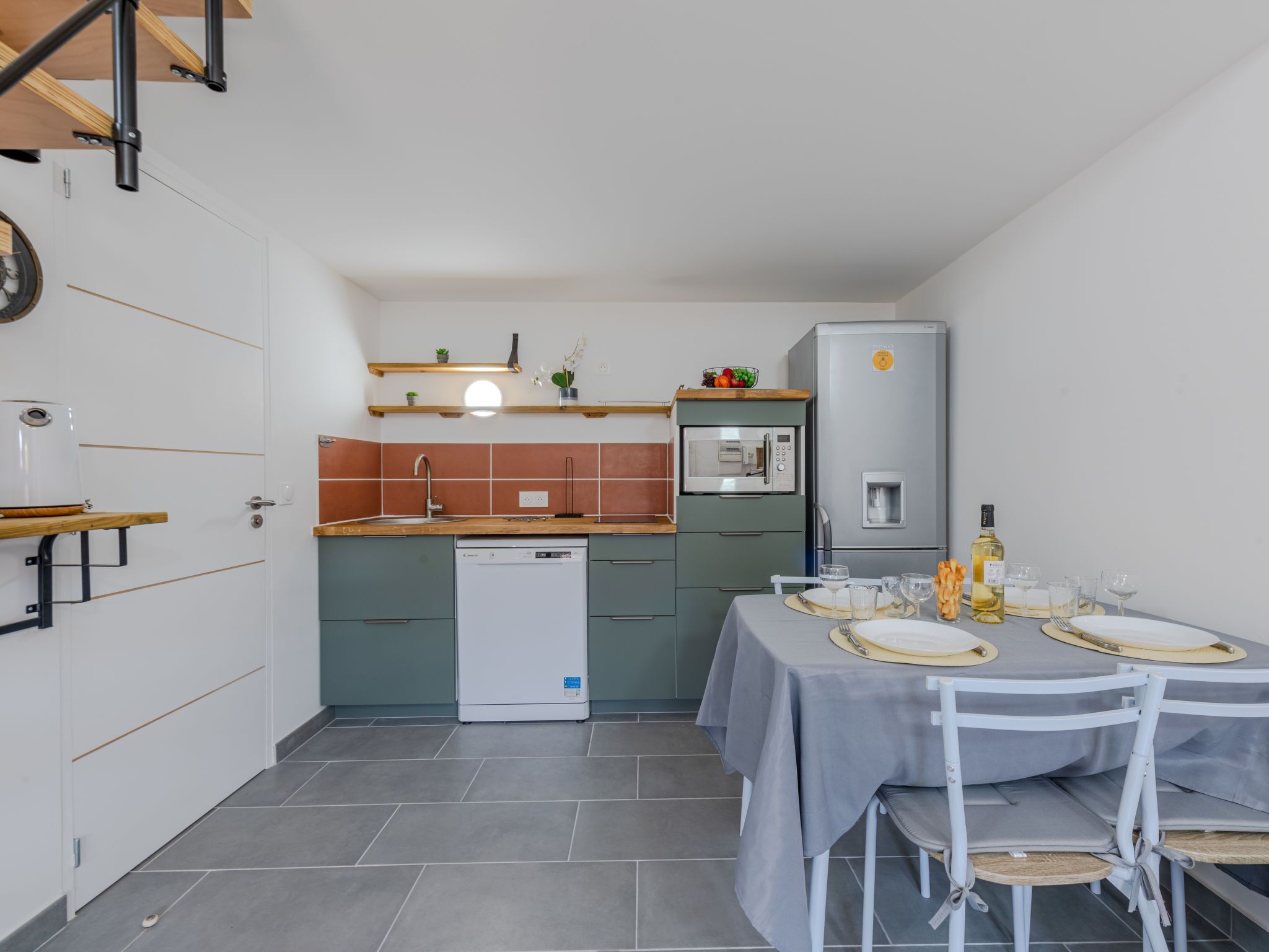 A compact kitchen with a dishwasher, microwave, and refrigerator. There is a dining table set with plates, glasses, and cutlery.