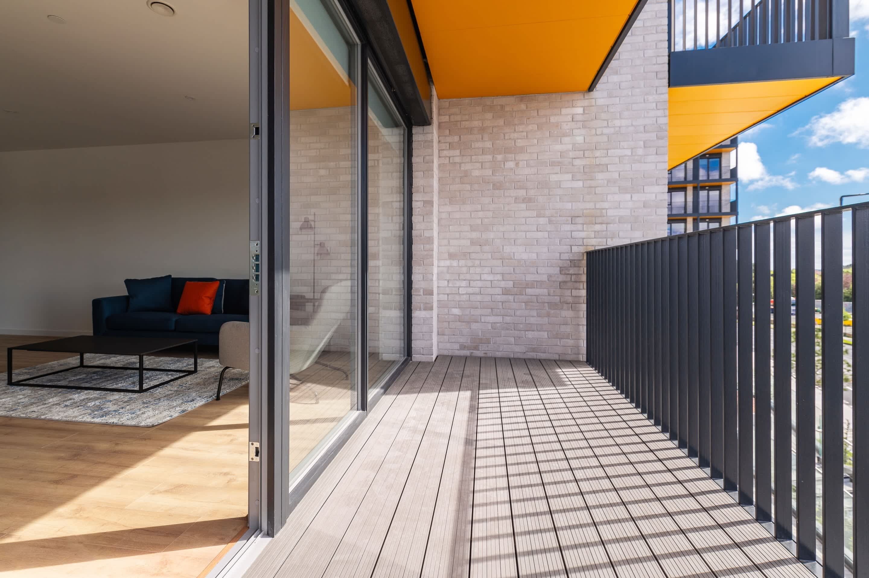 A balcony with a wooden floor, a black metal fence, and a glass door leading to an interior space with a sofa and a coffee table.
