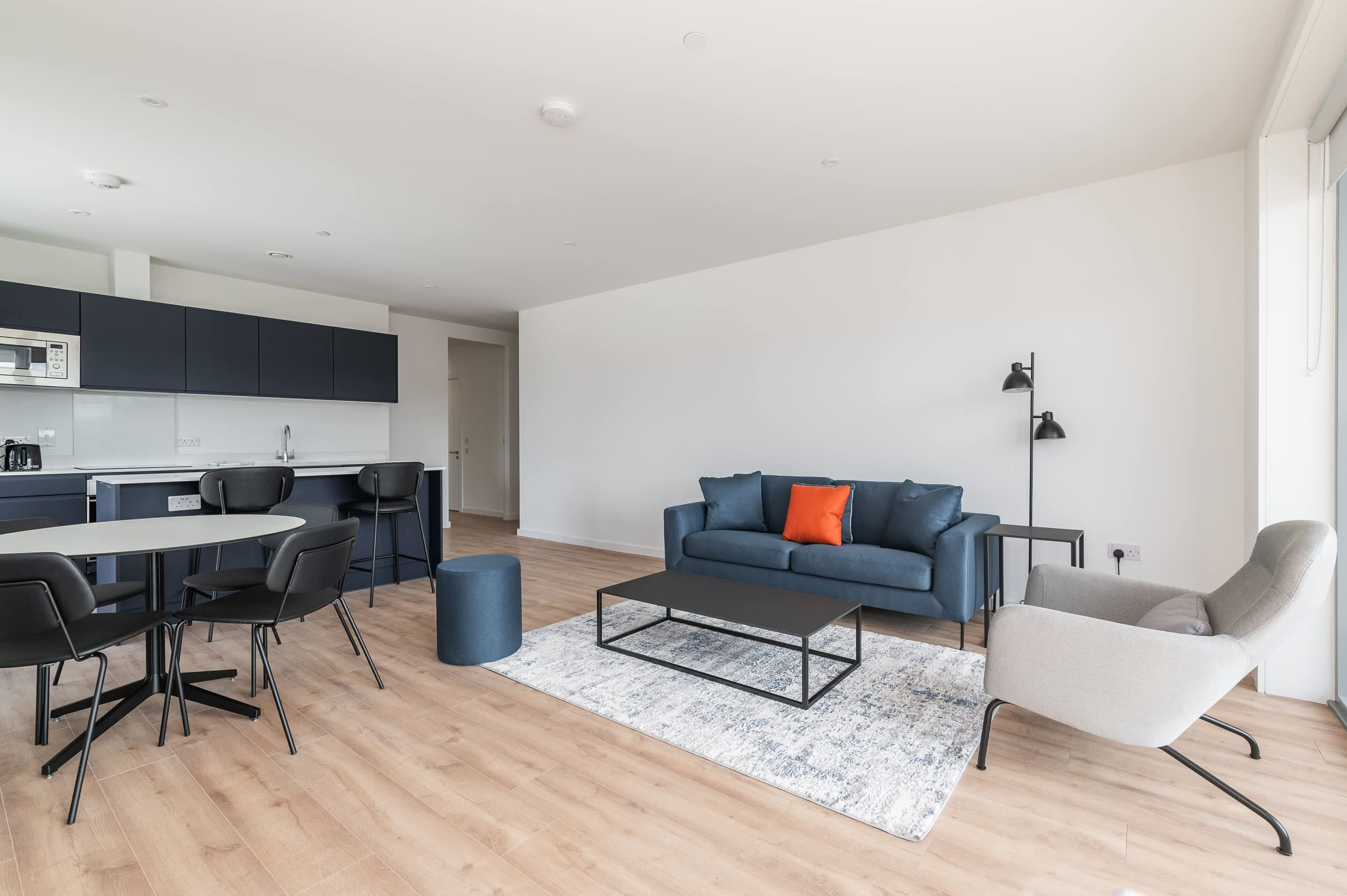Ein modernes Wohnzimmer mit einer Couch, einem Couchtisch und Sesseln. Im Hintergrund ist eine Küche mit Schränken und Geräten erkennbar.