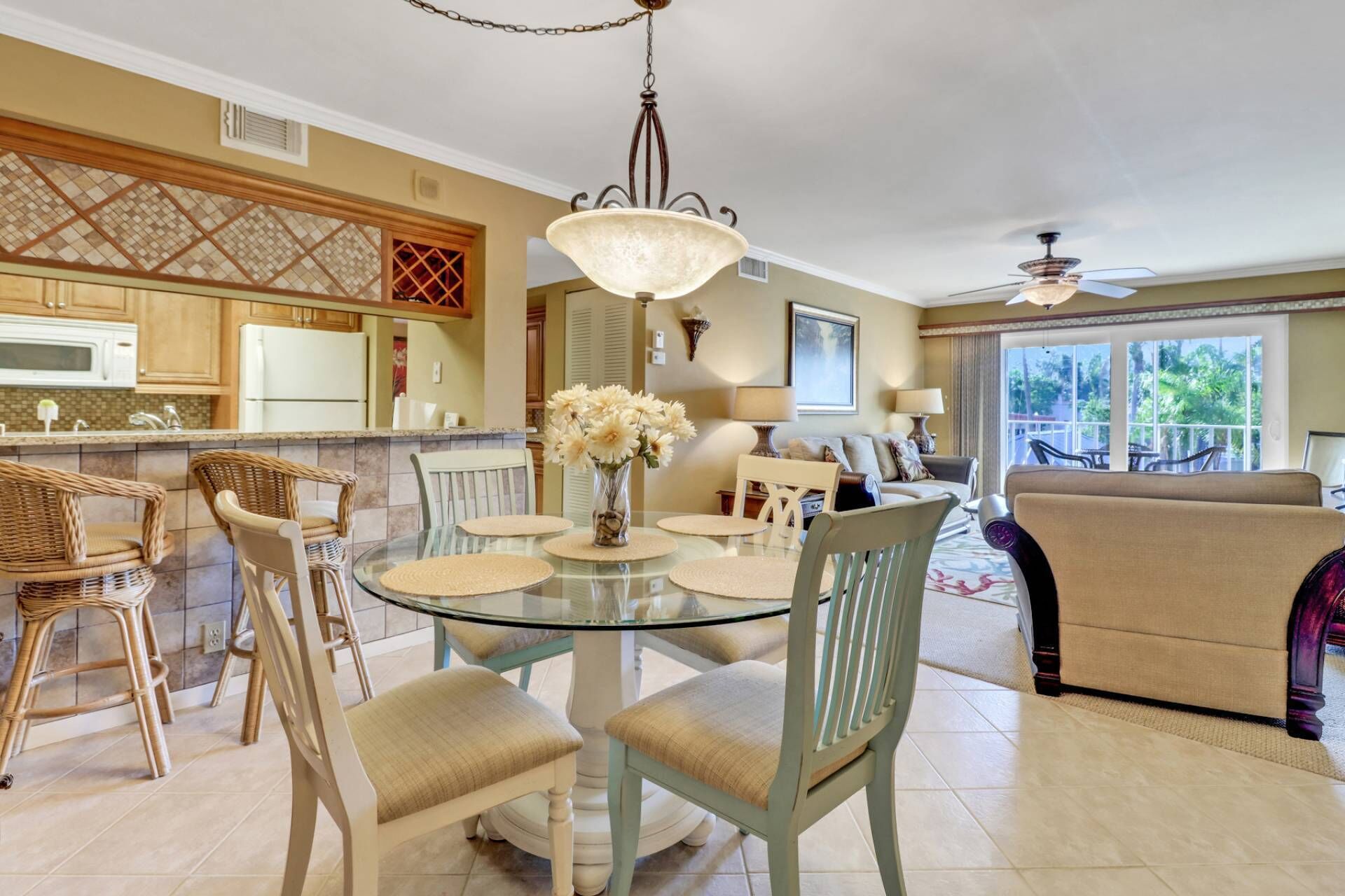 Uma sala de jantar com uma mesa de vidro rodeada por cadeiras de vime, um candelabro e vista para uma sala de estar com sofá e ventoinha de teto.