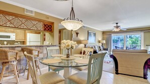 A dining area with a glass table surrounded by wicker chairs, a chandelier, and a view of a living room with a sofa and a ceiling fan. - Fort Myers River District Condo Close to Shopping, Dining & Nightlife! Heated Pool & Free Parking! (Fort Myers)