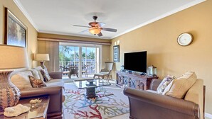A spacious living room with a ceiling fan, a flat-screen TV, and a large window overlooking a balcony with palm trees. - Fort Myers River District Condo Close to Shopping, Dining & Nightlife! Heated Pool & Free Parking! (Fort Myers)