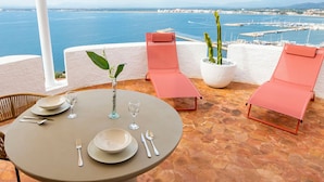 A terrace with a table set for two, two pink lounge chairs, a potted plant, and a view of the sea and marina. - Castell Mar 1007 in Roses (Roses)