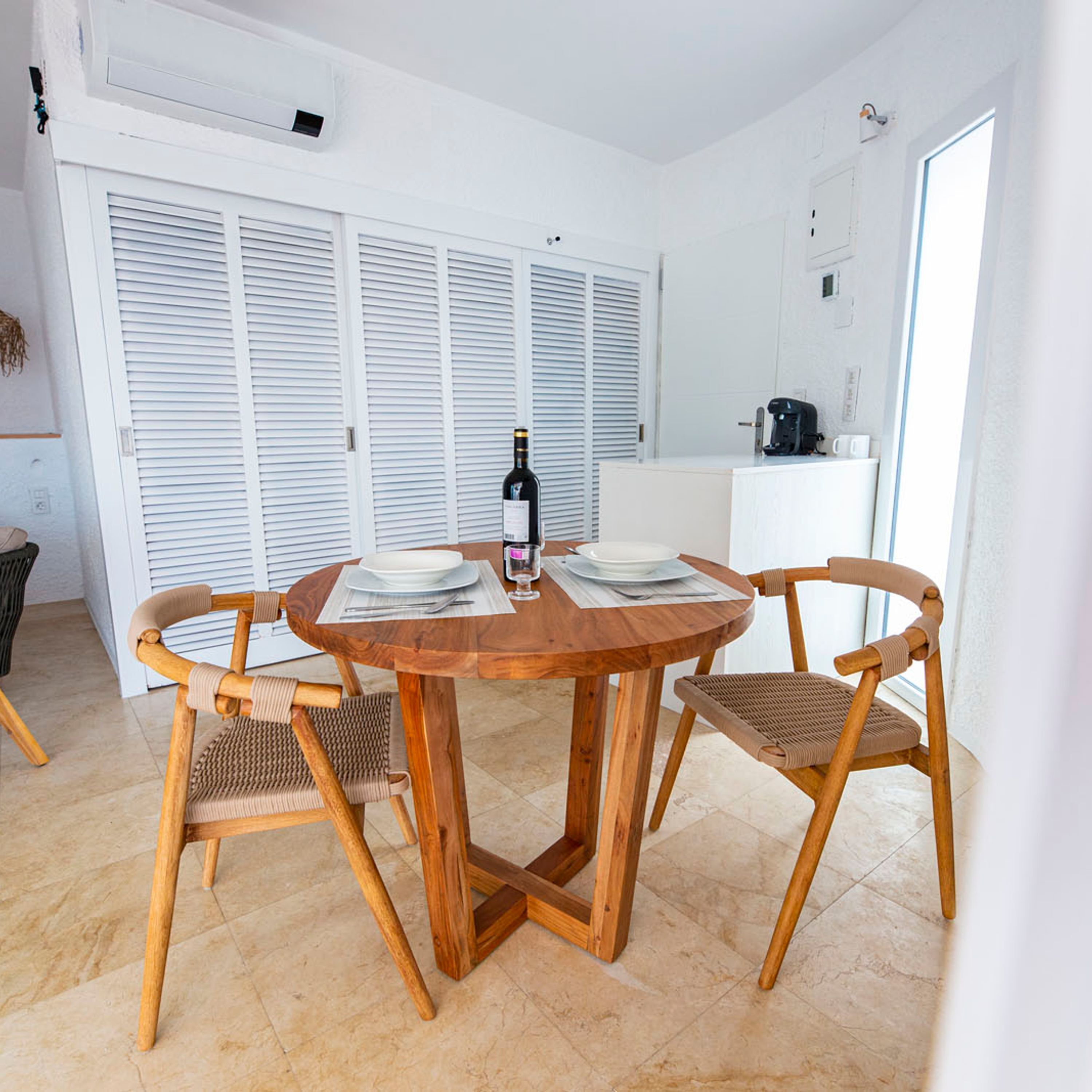 A dining area with a round wooden table, two wicker chairs, and a bottle of wine.