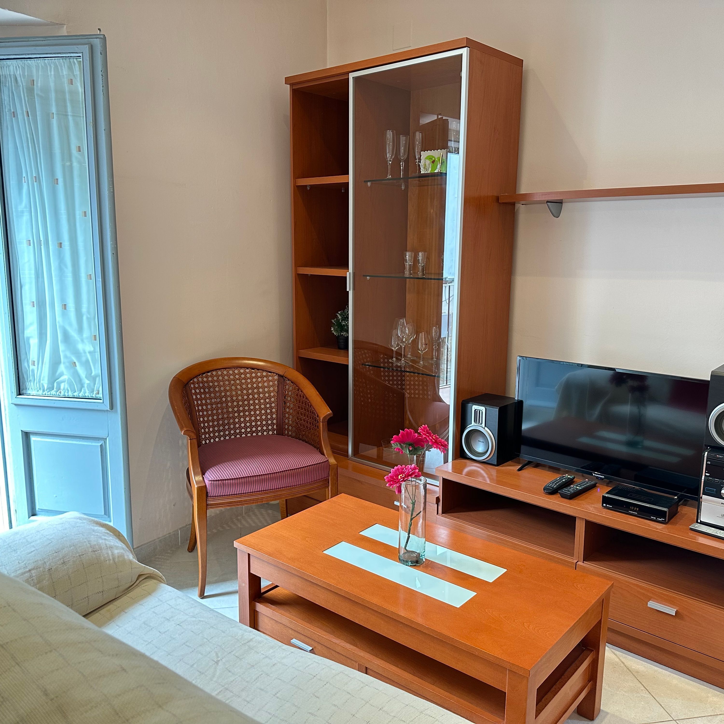 A room with a wooden TV stand, a flat-screen TV, a wooden cabinet with shelves, a wicker chair, and a small table with a vase of flowers.