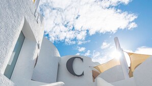 Um edifício branco com um 'C' grande na fachada, uma escadaria que leva a uma porta e um céu azul claro ao fundo.