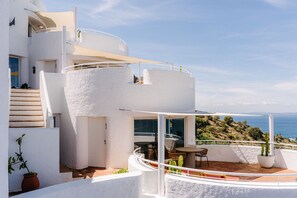 A modern white building with a curved balcony, outdoor seating, and a view of the sea.