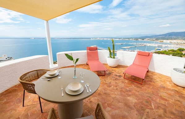 A terrace with a table set for two, two chairs, a potted plant, and a view of the sea and marina.