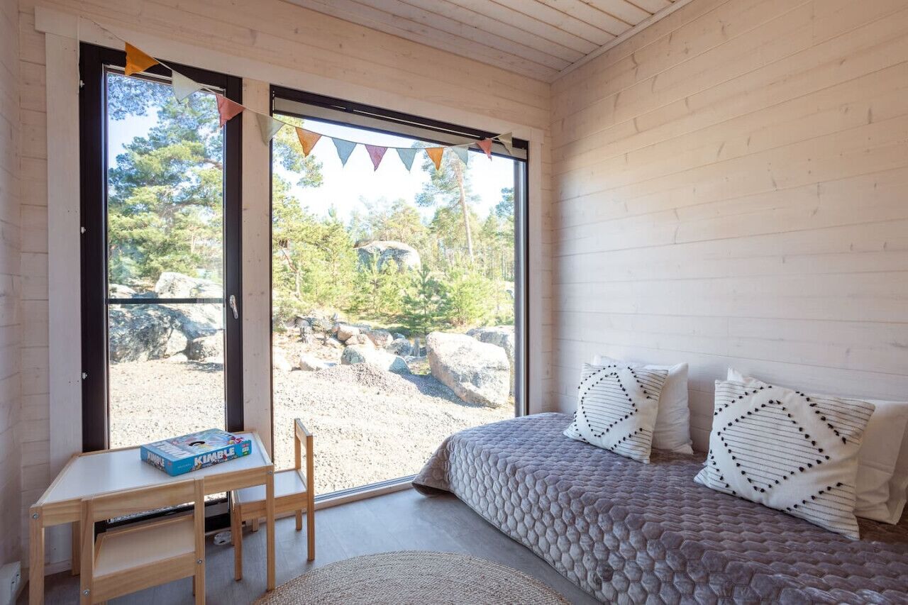 Une chambre avec une grande porte-fenêtre coulissante donnant sur un espace extérieur, une table en bois avec un jeu de société, un lit avec des coussins à motifs et un tapis tissé.