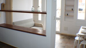 A room with a wooden counter, a white chair, and a dining table with chairs. There are shelves with decorative items and a window providing natural light. - Porthos 113 in Roses (Roses)