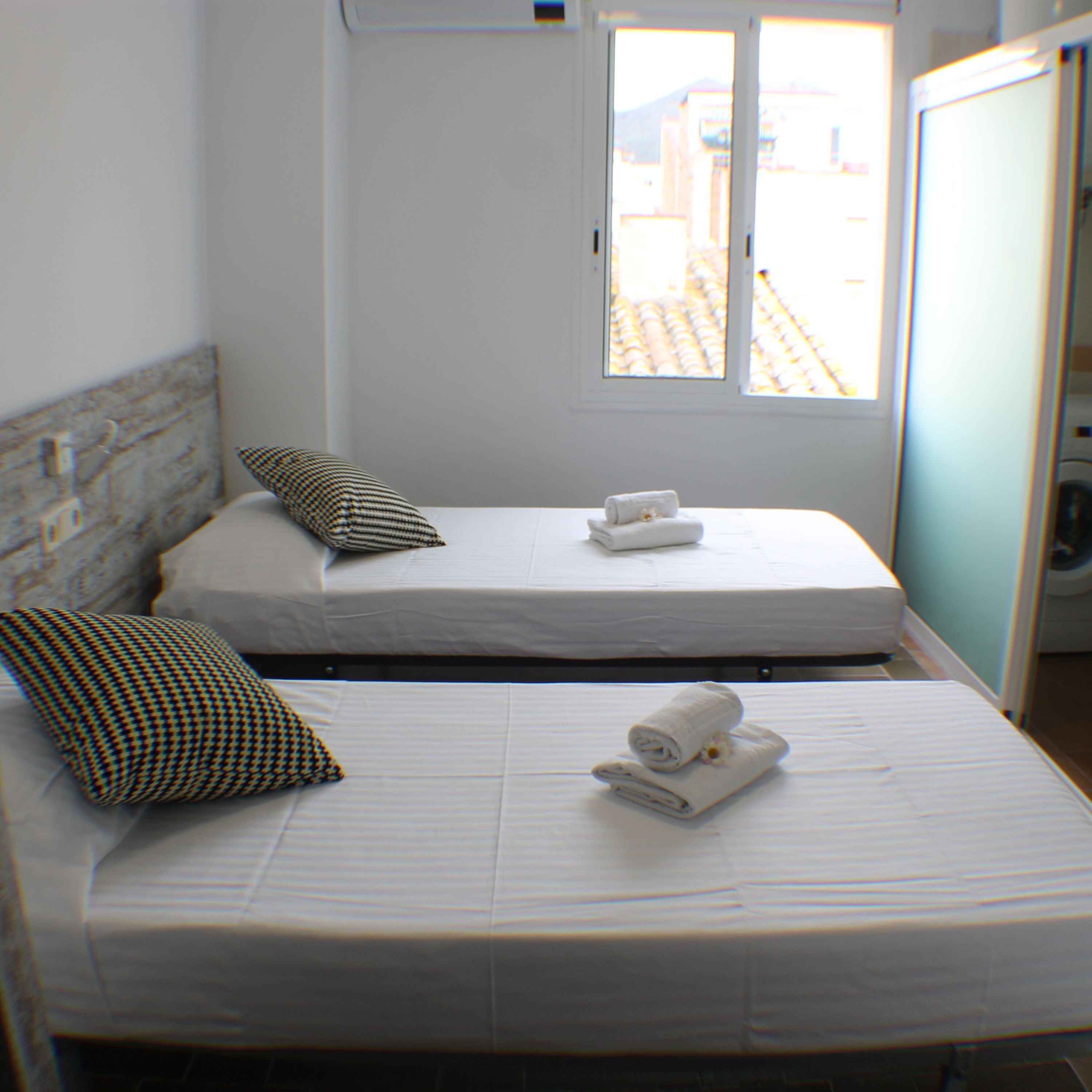 A hotel room with two beds, each with a pillow and rolled towels. There is a window with a view of buildings outside.
