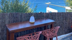 Una mesa de madera con dos sillas, un altavoz blanco y vistas al mar y un paisaje urbano distante.