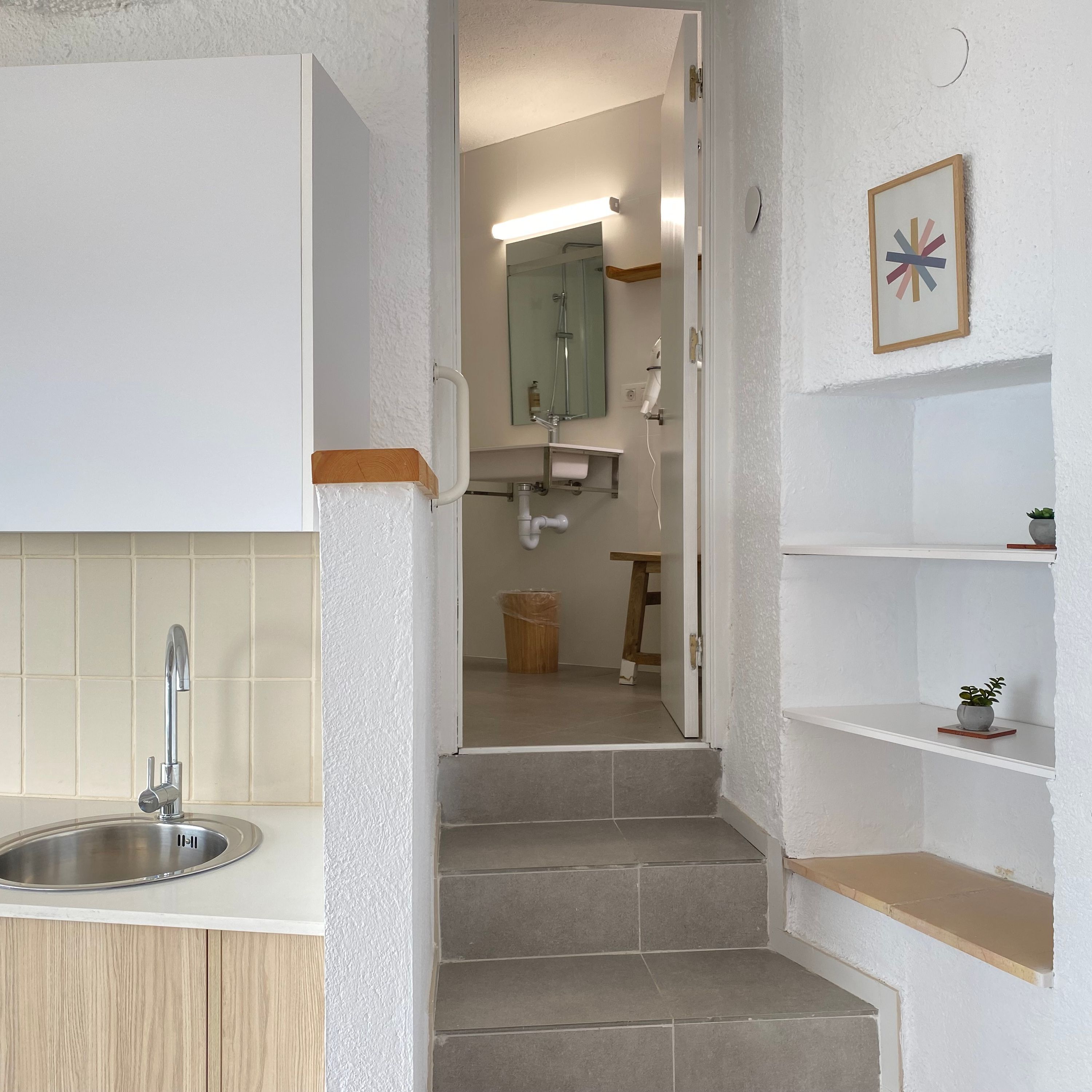 Un baño moderno con lavamanos, espejo y ducha. Hay escalones que conducen a la zona de la ducha, y una pequeña estantería con una planta.